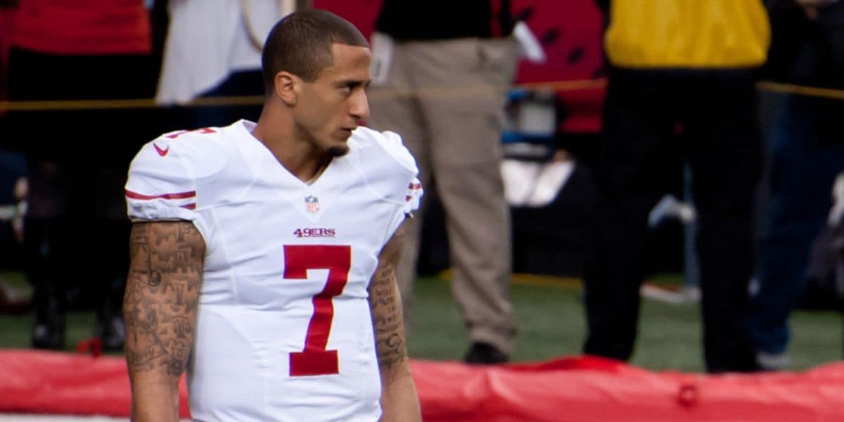 A football player wearing a white San Francisco 49ers uniform with the number 7 stands on the field, looking to the side. He has short hair and visible tattoos on both arms. The background shows blurred people and equipment.