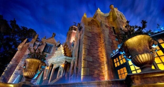 A dramatic, low-angle view of a gothic-style brick mansion at night, illuminated with eerie blue and yellow lights, with large stone urns and intricate architectural details visible in the foreground.