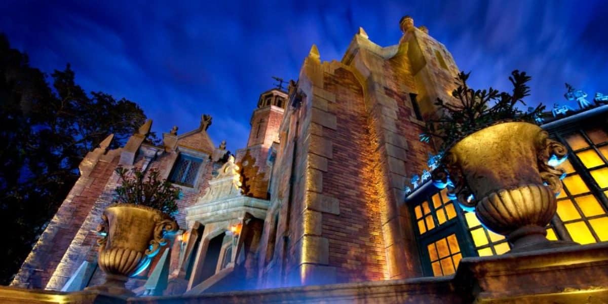 A dramatic, low-angle view of a gothic-style brick mansion at night, illuminated with eerie blue and yellow lights, with large stone urns and intricate architectural details visible in the foreground.
