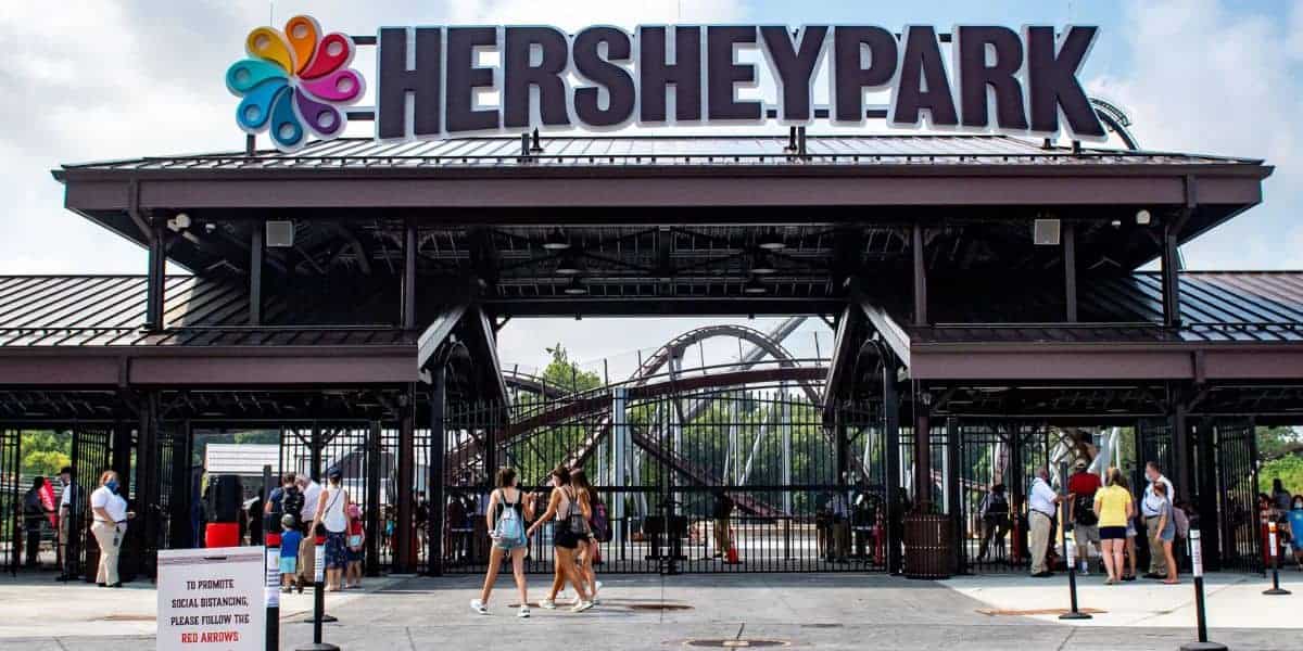 Entrance to Hersheypark with a large sign overhead, people walking in and out, a roller coaster visible in the background, and a sign with entry instructions in the foreground.