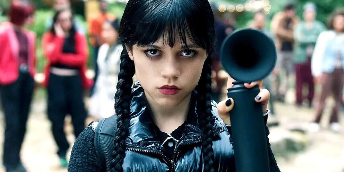 A girl with long black braided hair and dark clothing stares intensely at the camera while holding up a black object, with a blurred group of people standing in the background outdoors.