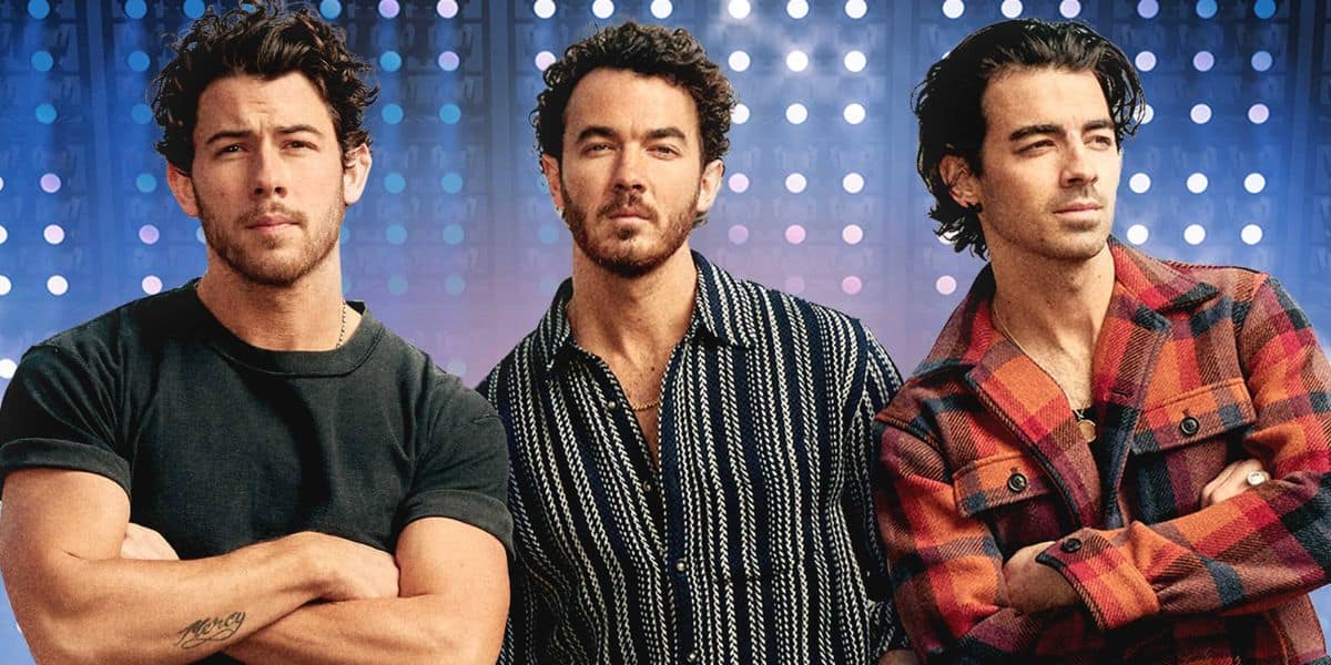 Three men with short, curly dark hair stand side by side against a backdrop of blue and purple lights. The men wear casual shirts and have neutral expressions, looking slightly off-camera.