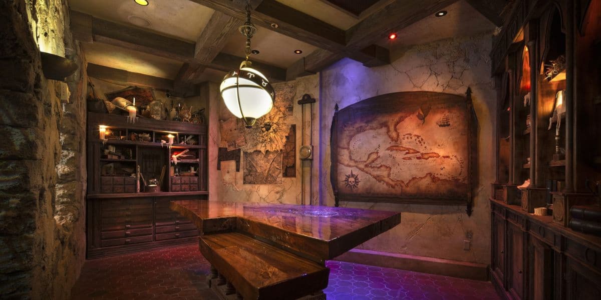 A dimly lit, rustic room with wooden beams, a large antique map on the wall, a triangular wooden table, shelves filled with artifacts, and a round hanging lantern. The decor evokes a vintage explorer or pirate theme.