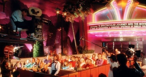 A group of people sits in a ride vehicle facing a large movie camera and film crew, with bright neon lights and a marquee reading "A Spectacular Journey Into The Movies" in the background.
