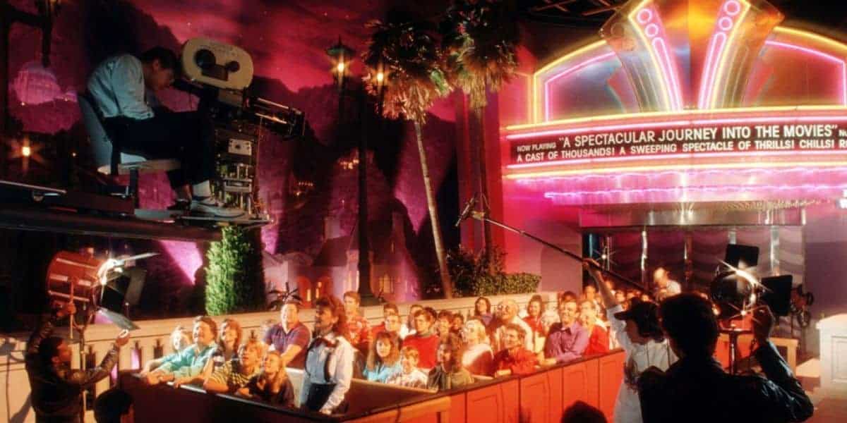 A group of people sits in a ride vehicle facing a large movie camera and film crew, with bright neon lights and a marquee reading "A Spectacular Journey Into The Movies" in the background.
