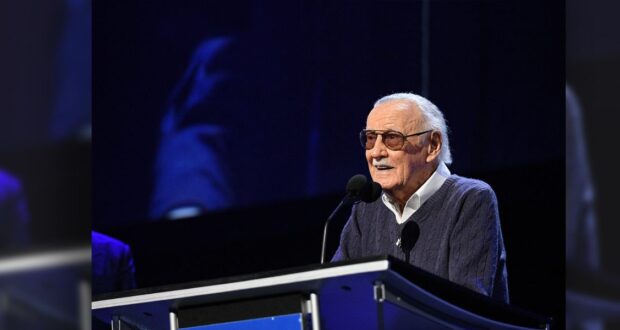 An elderly man wearing sunglasses and a sweater speaks at a podium with a sign that reads "D23 Expo: The Ultimate Disney Fan Event." The background is dark and the scene is brightly lit.