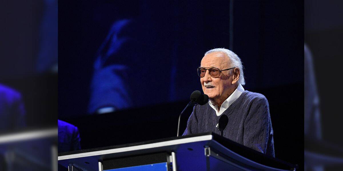 An elderly man wearing sunglasses and a sweater speaks at a podium with a sign that reads "D23 Expo: The Ultimate Disney Fan Event." The background is dark and the scene is brightly lit.