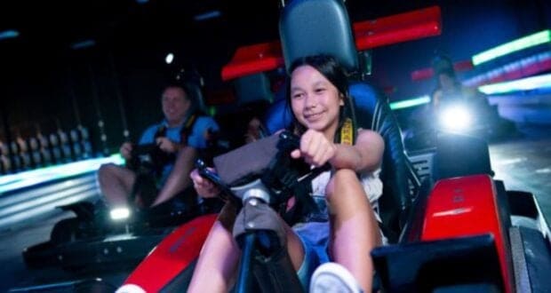 A young girl smiles while driving a red go-kart indoors, gripping the steering wheel. Other racers and colorful neon lights are visible in the background.