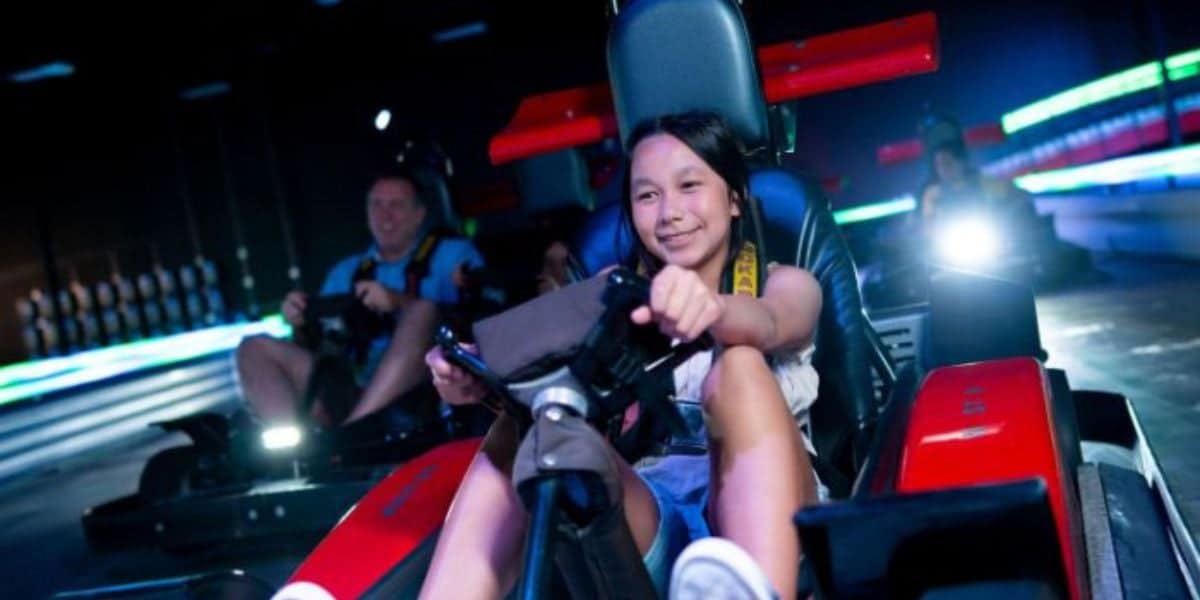 A young girl smiles while driving a red go-kart indoors, gripping the steering wheel. Other racers and colorful neon lights are visible in the background.