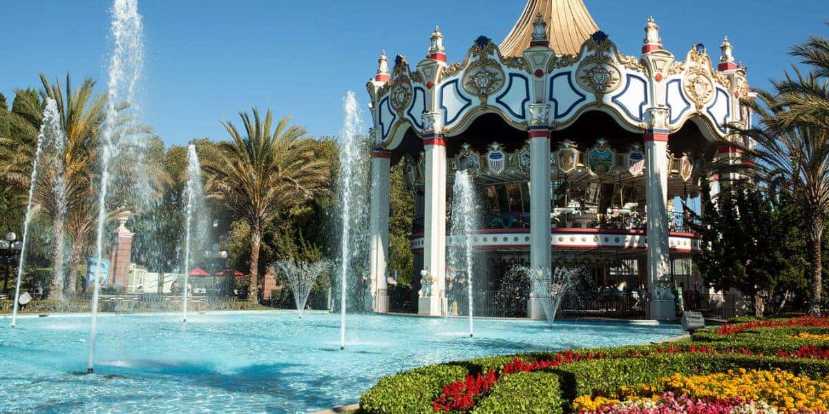 A colorful carousel with ornate decorations stands behind a large, clear fountain surrounded by palm trees, flowers, and neatly manicured greenery under a bright blue sky.