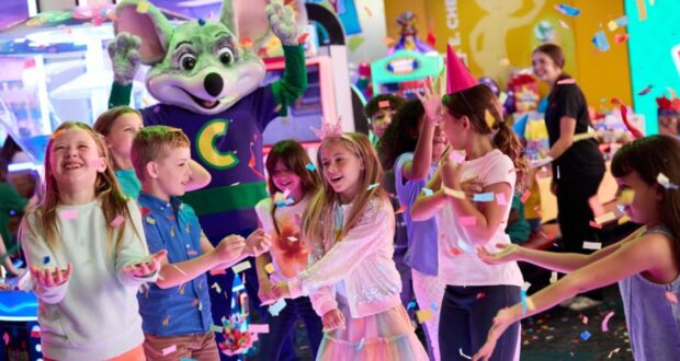 A group of children at a birthday party celebrate with confetti, smiling and laughing. A person in a large mouse mascot costume stands behind them with raised arms. Bright arcade games and decorations fill the colorful background.