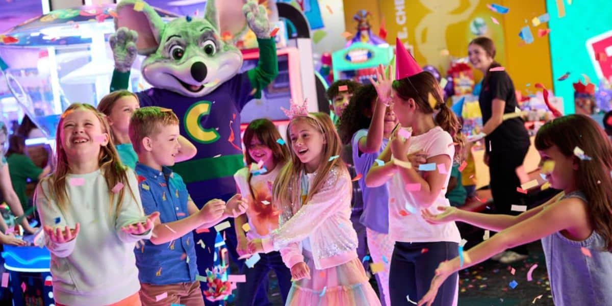 A group of children at a birthday party celebrate with confetti, smiling and laughing. A person in a large mouse mascot costume stands behind them with raised arms. Bright arcade games and decorations fill the colorful background.