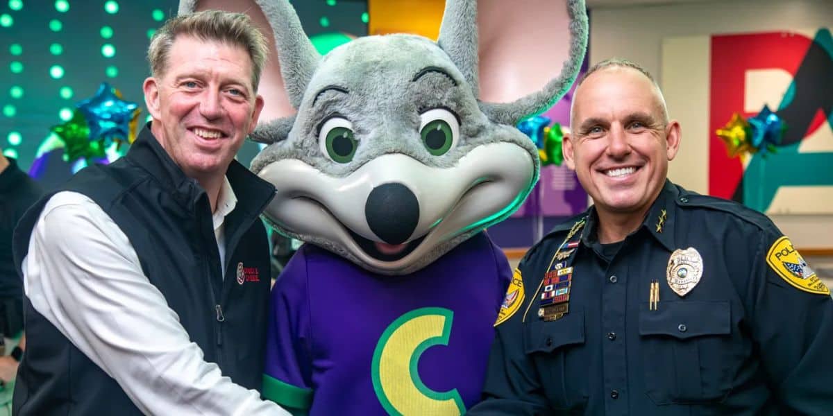 Two men, one in a business vest and one in a police uniform, pose and smile on either side of a person in a large gray mouse mascot costume wearing a purple shirt with a yellow "C" on it.