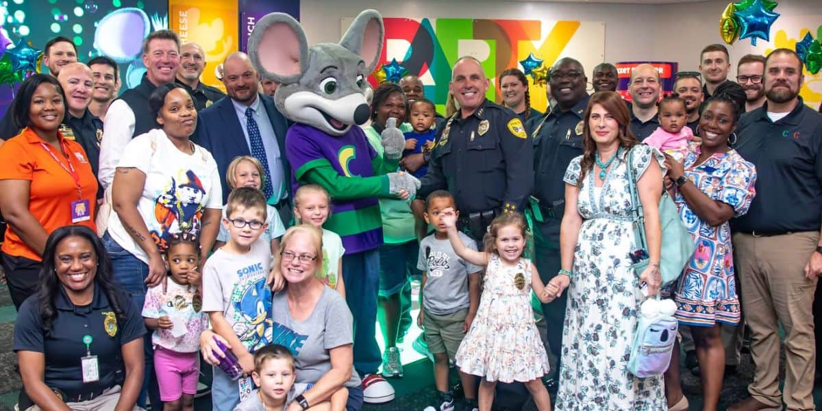 A large, diverse group of adults and children, including police officers and a person in a mouse mascot costume, pose and smile together at a colorful indoor party venue with balloons and decorations.