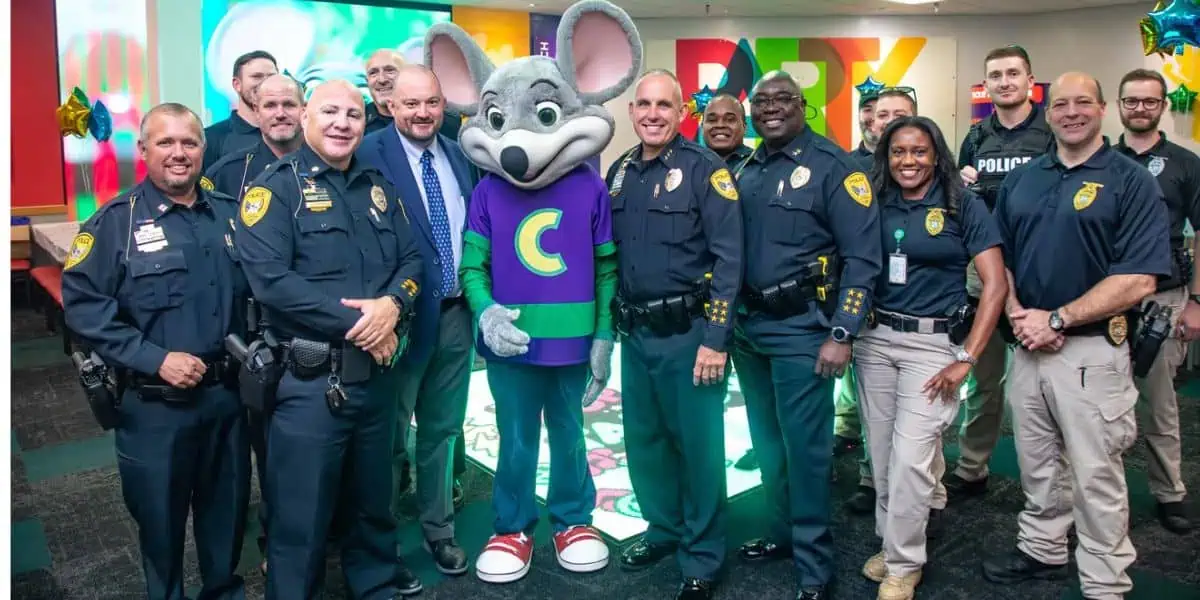 A group of police officers and staff smile and pose for a photo with the Chuck E. Cheese mascot inside a colorful, decorated indoor party venue.