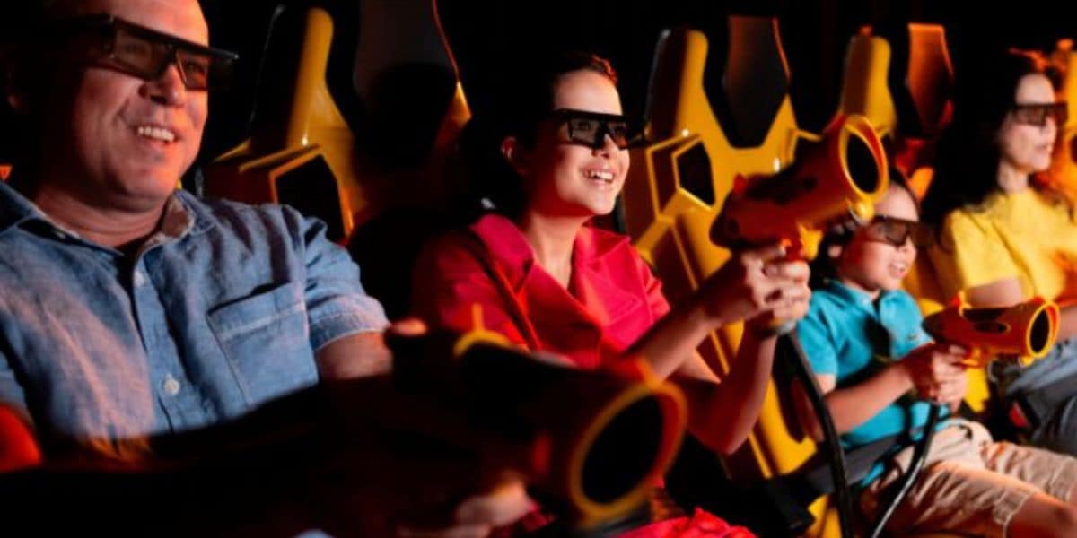 Four people wearing 3D glasses sit in yellow seats, smiling and holding game controllers, as if enjoying an interactive ride or virtual reality experience. Warm lighting highlights their faces.
