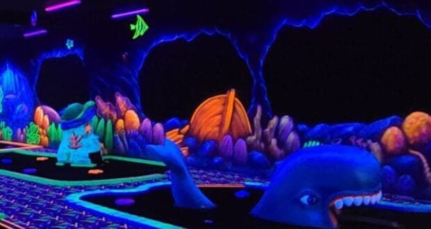 A glow-in-the-dark mini-golf course with an underwater theme, featuring colorful sea creatures, coral, and a large whale sculpture under black lights. Neon colors and sea decorations line the walls and course.