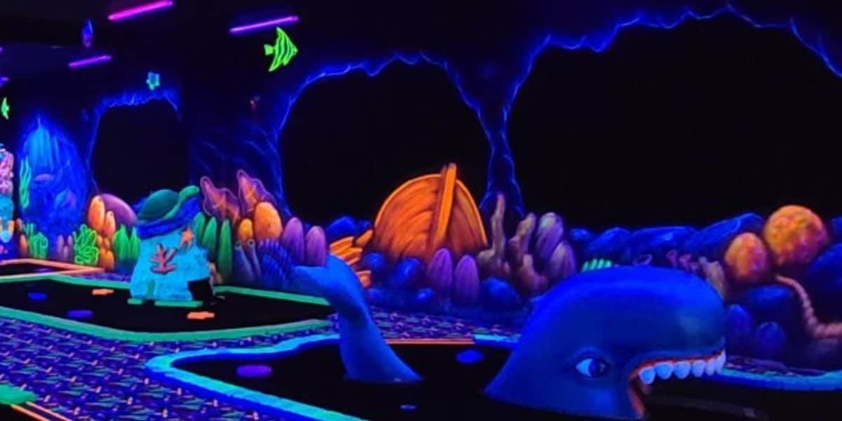 A glow-in-the-dark mini-golf course with an underwater theme, featuring colorful sea creatures, coral, and a large whale sculpture under black lights. Neon colors and sea decorations line the walls and course.