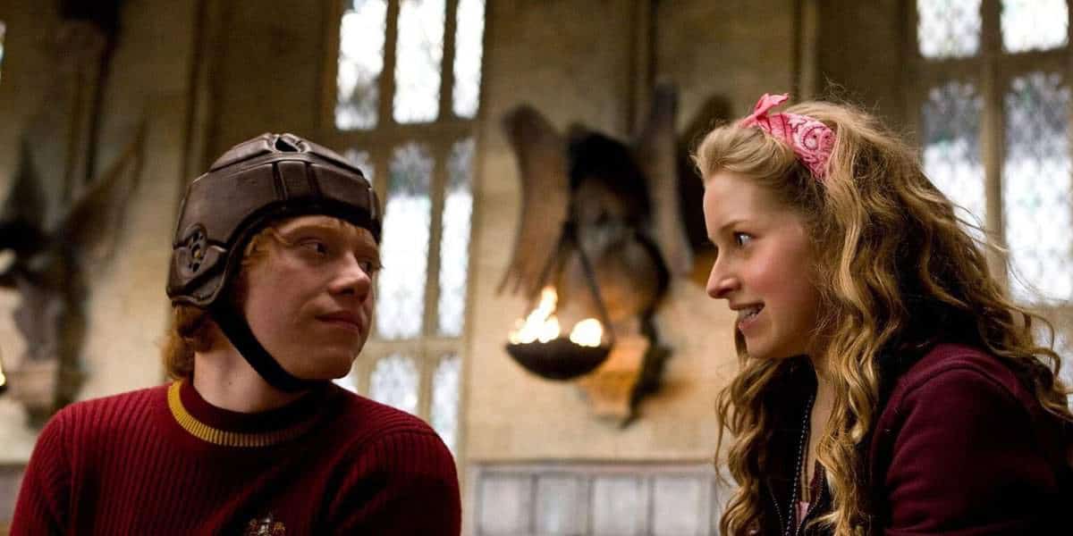 A young man in a brown Quidditch helmet and red sweater looks at a smiling young woman with long curly hair and a pink headband, sitting together in a medieval-style hall with stone walls and arched windows.