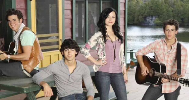 Four young people stand and sit on a wooden deck by a lake, with two holding guitars. A cabin with green trim and a yellow door is behind them, and trees are in the background.