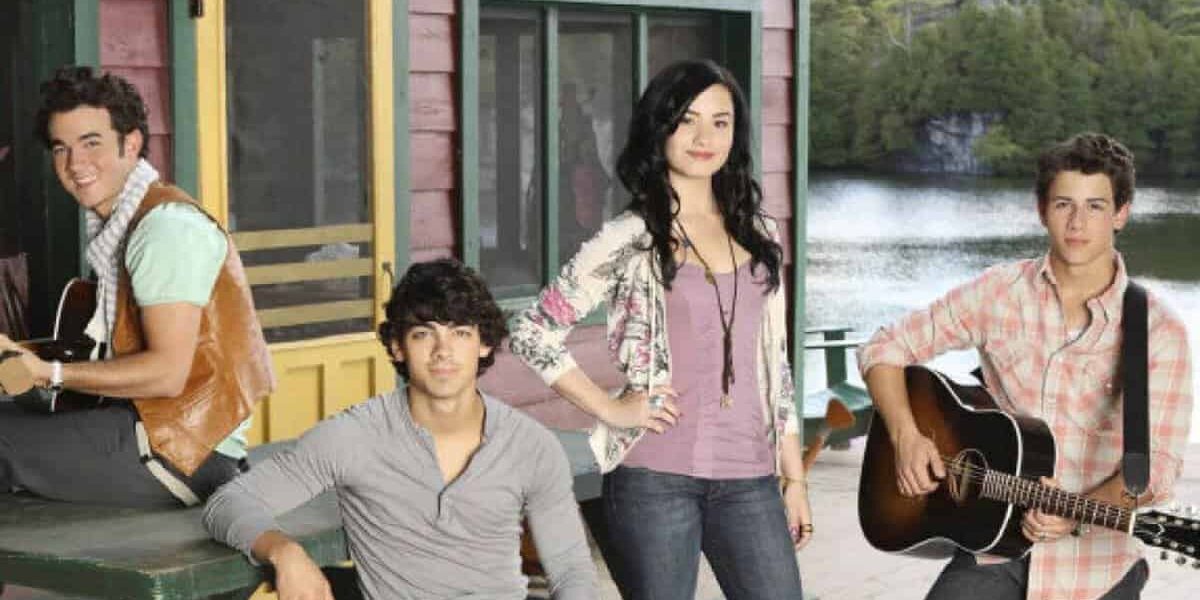 Four young people stand and sit on a wooden deck by a lake, with two holding guitars. A cabin with green trim and a yellow door is behind them, and trees are in the background.