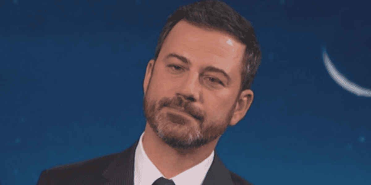 A man with short dark hair and a beard, wearing a suit and tie, stands against a dark blue background with a crescent moon visible. He has a serious, slightly skeptical expression.