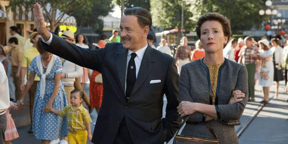 A man in a suit cheerfully waves while a woman beside him stands with her arms crossed, looking unimpressed. They are outside, surrounded by people walking in a lively, sunny setting.