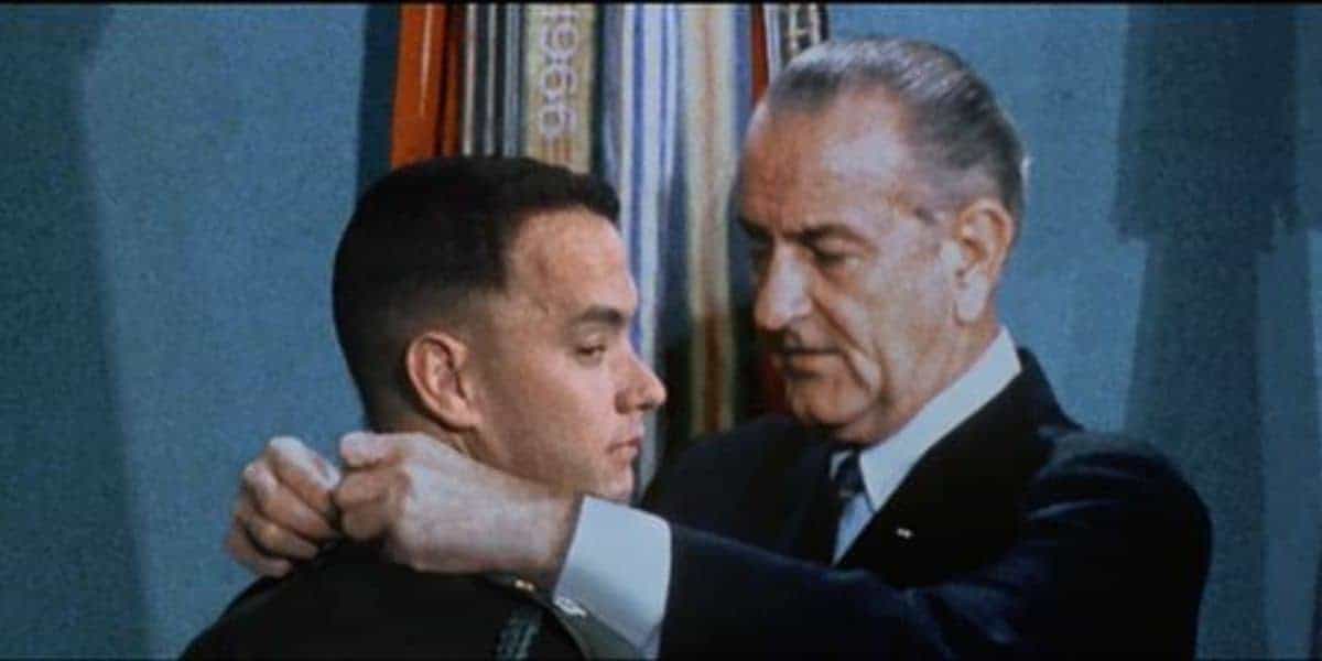 A man in a military uniform stands as an older man in a suit places a medal around his neck, with flags in the background.