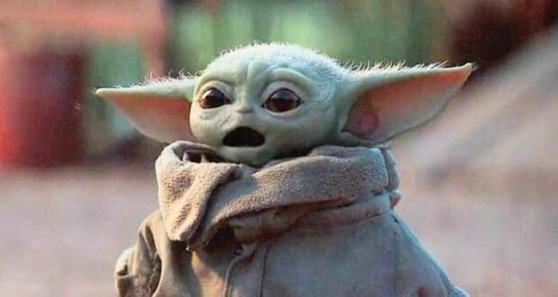 A small, green, big-eared creature known as Baby Yoda or Grogu, wearing a brown robe and scarf, stands outdoors with a surprised expression.
