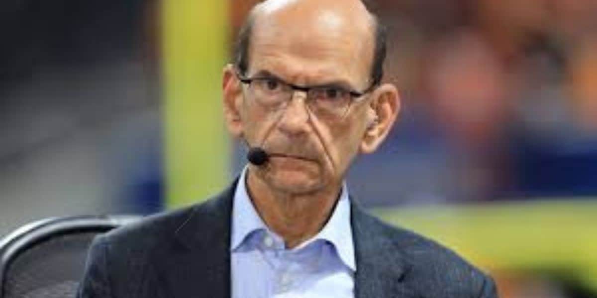 A man wearing glasses and a suit jacket sits with a serious expression, wearing a headset microphone, against a blurred background.