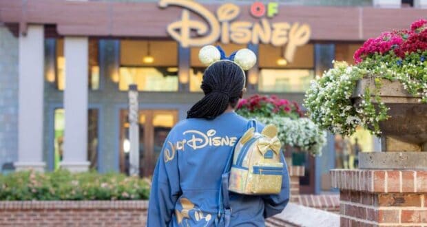 A person wearing Mickey Mouse ears and a blue Disney jacket with a matching backpack stands outside a Disney store, facing the store’s entrance. Bright flowers and the Disney sign are visible in the background.