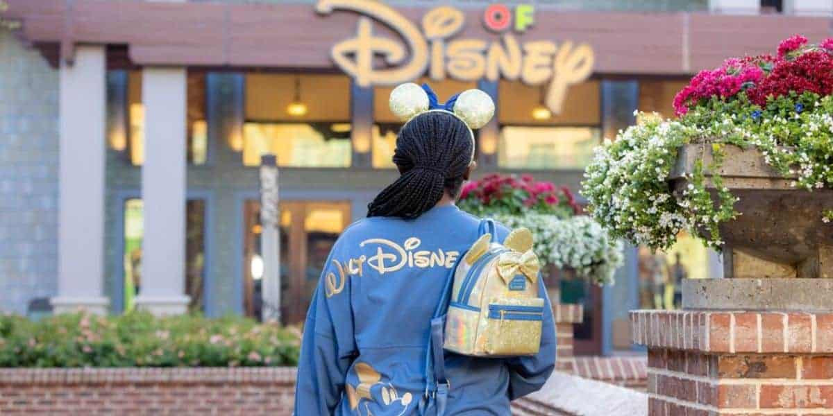 A person wearing Mickey Mouse ears and a blue Disney jacket with a matching backpack stands outside a Disney store, facing the store’s entrance. Bright flowers and the Disney sign are visible in the background.