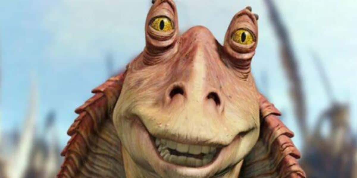 A close-up of Jar Jar Binks, a fictional alien character with yellow eyes and a wide smile, from the Star Wars movie series, against a blurred outdoor background.