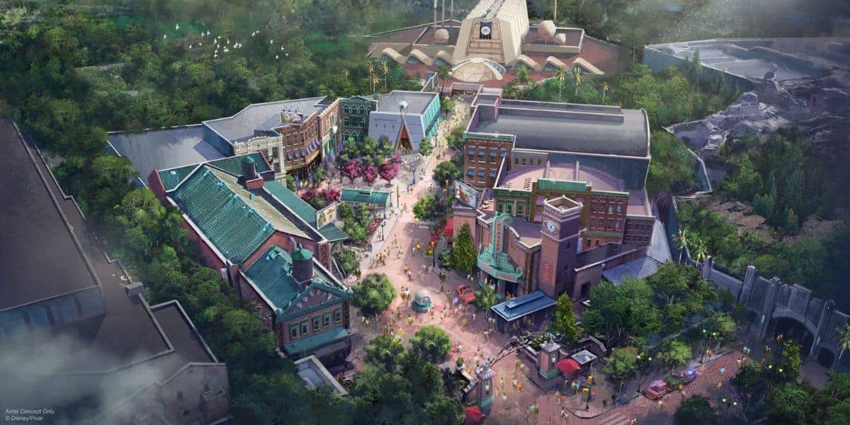 Aerial illustration of a theme park area with colorful buildings, tree-lined streets, pathways, a central fountain, and people walking. In the background, large structures and greenery surround the vibrant scene.