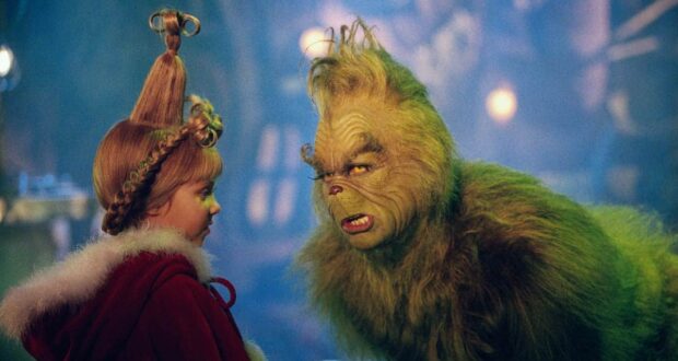 A young girl with tall, styled hair in a red cloak faces a furry green creature with a grumpy expression in a festive, whimsical setting.