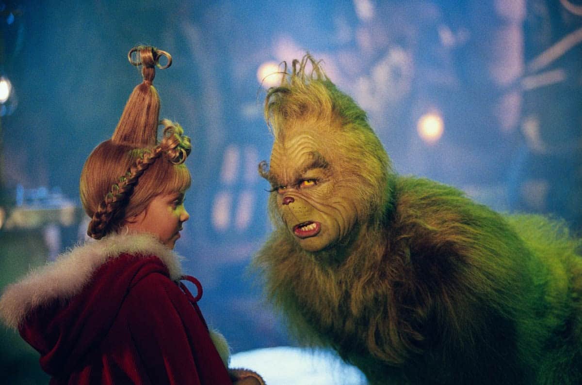 A young girl with tall, styled hair in a red cloak faces a furry green creature with a grumpy expression in a festive, whimsical setting.