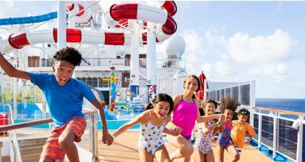 A group of smiling children and one adult in swimsuits run excitedly on a cruise ship deck near a large water park with slides, under a blue sky.