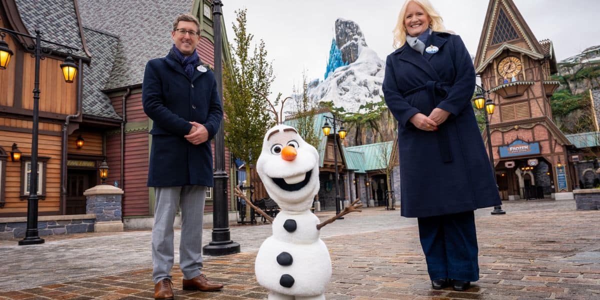 Guests in dark coats pose with Interactive Olaf from Frozen at a snowy Disney park area crafted by Walt Disney Imagineering.