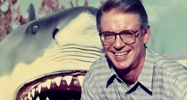 A smiling man wearing glasses and a patterned shirt stands in front of a large, open-mouthed shark model, possibly from a movie set or theme park attraction.