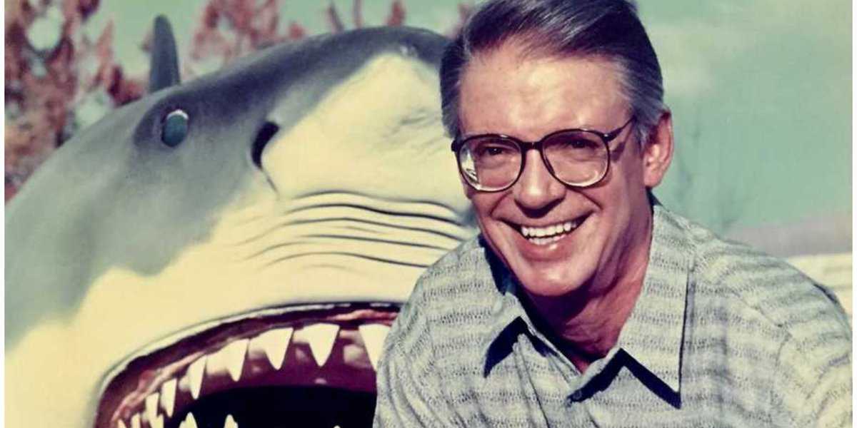 A smiling man wearing glasses and a patterned shirt stands in front of a large, open-mouthed shark model, possibly from a movie set or theme park attraction.