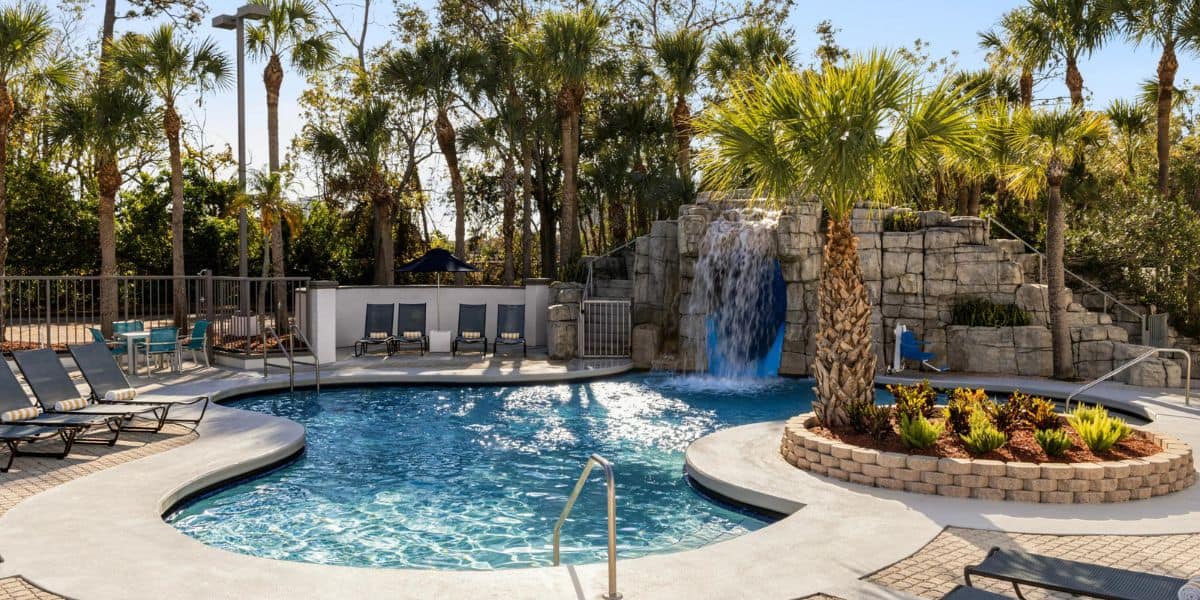 Outdoor swimming pool with lounge chairs, palm trees, and a rock waterfall feature under a bright sky. There is a small island with plants in the pool and a fenced seating area to the left.