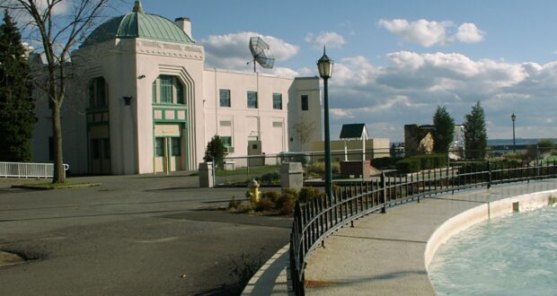 A white building with a green dome stands near a landscaped walkway, lampposts, and a curved, partially-filled fountain. The area is under a cloudy sky, with trees and water visible in the background.