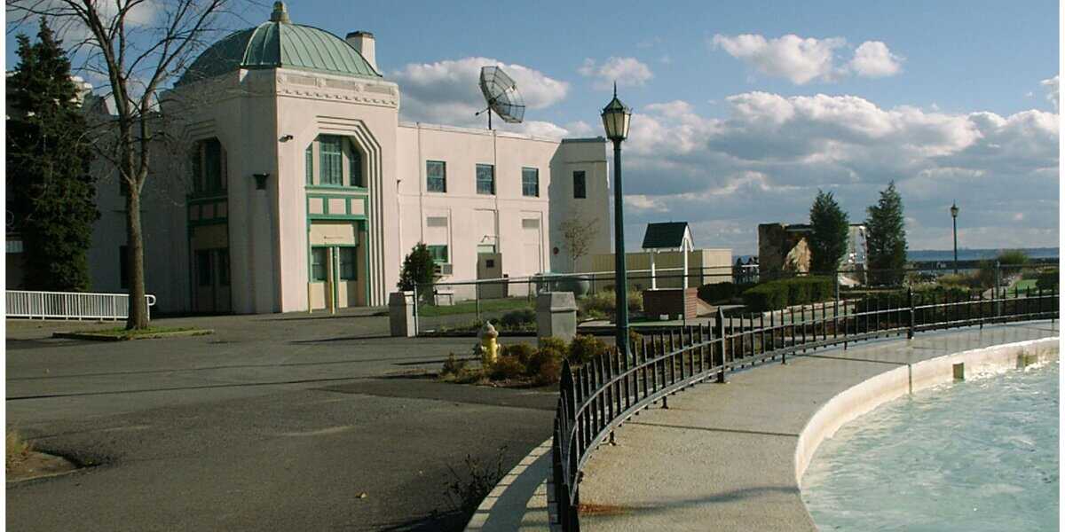 A white building with a green dome stands near a landscaped walkway, lampposts, and a curved, partially-filled fountain. The area is under a cloudy sky, with trees and water visible in the background.