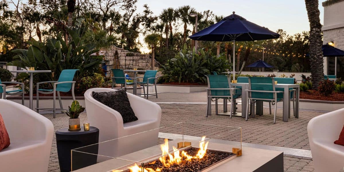 Outdoor patio with modern seating around a rectangular fire pit, teal chairs at tables with navy umbrellas, lush green plants in the background, and string lights at sunset.