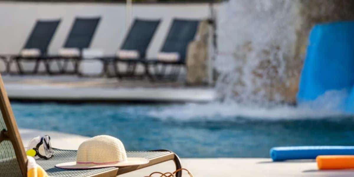 A sun hat, sunglasses, and a towel rest on a lounge chair by a swimming pool, with water splashing from a fountain and empty lounge chairs in the background.