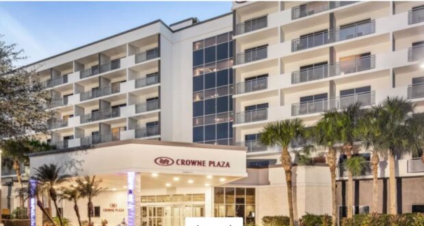 Exterior view of a multi-story Crowne Plaza hotel with balconies, glass windows, palm trees, and a well-lit entrance canopy at dusk.