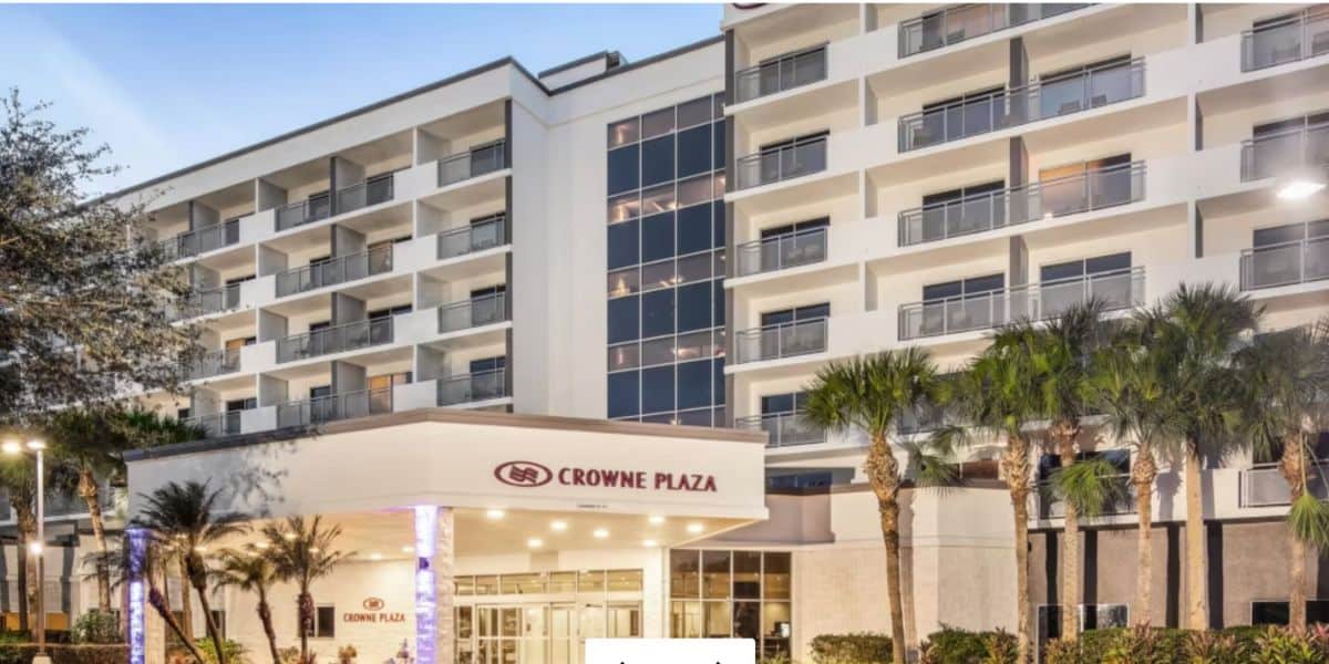 Exterior view of a multi-story Crowne Plaza hotel with balconies, glass windows, palm trees, and a well-lit entrance canopy at dusk.