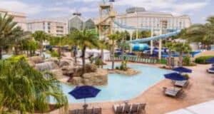 Thrilling resort pool zone with palm trees, winding water slides, lounge seating, shaded umbrellas, and towering hotel backdrops.