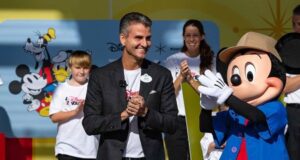 Mickey Mouse and an executive share applause on stage with Disney fans, set against a vibrant Disney World backdrop.