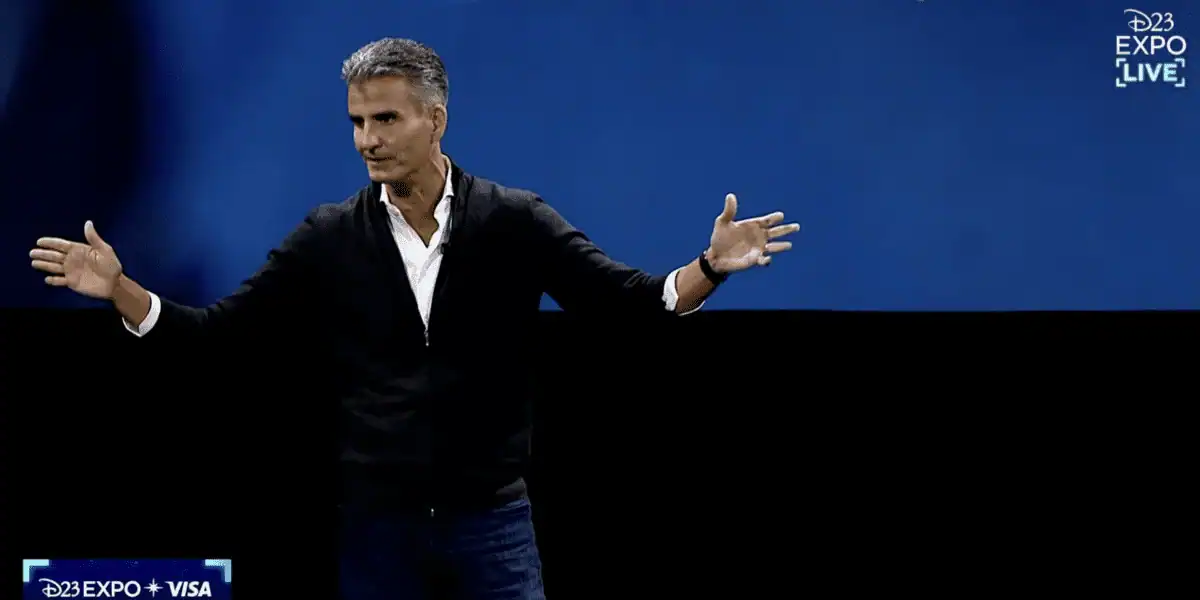 A Disney presenter in a black sweater speaks onstage at D23 Expo, set against a vibrant blue backdrop.